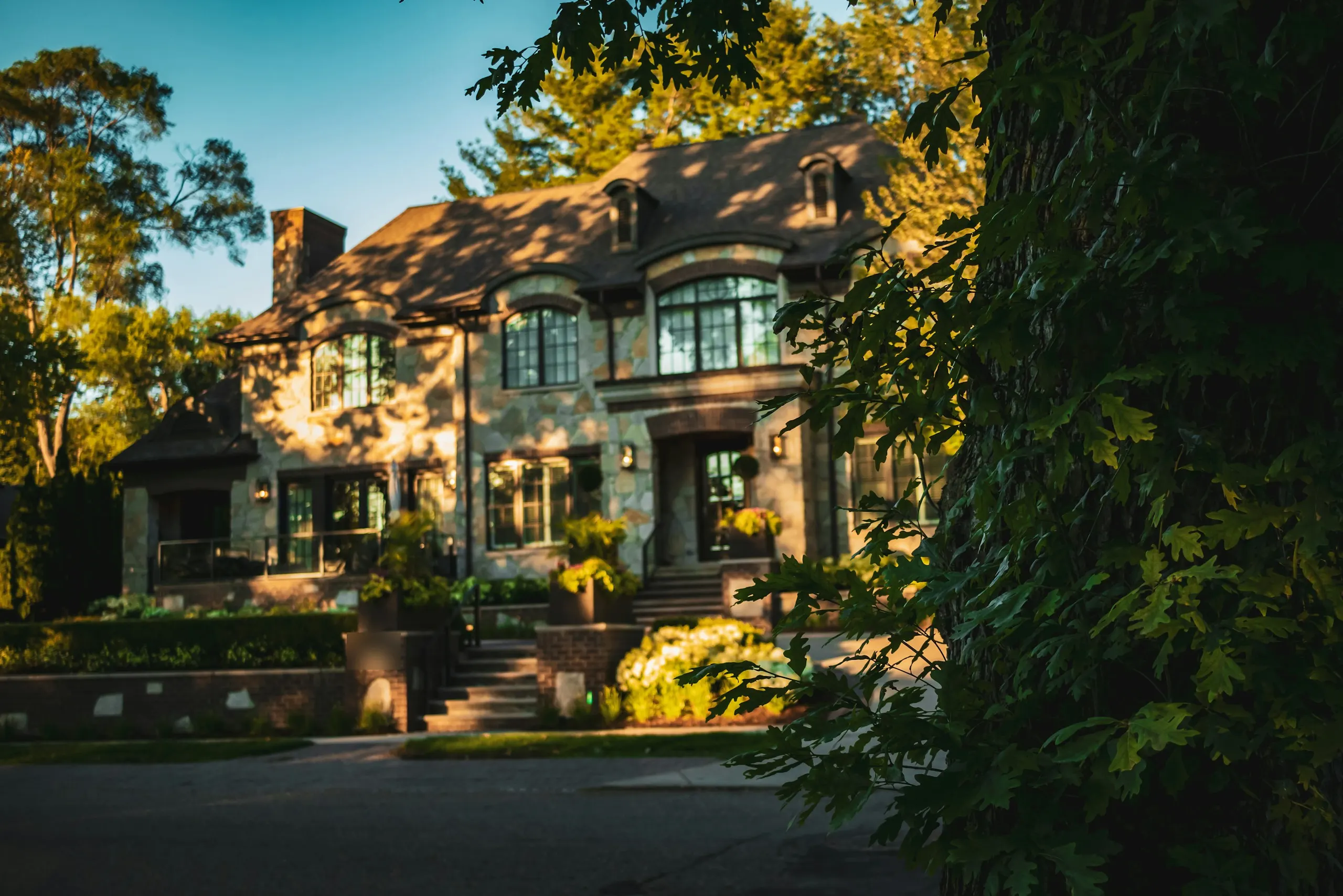 A stone and brick family home glowing in late-afternoon sun behind a mature oak
